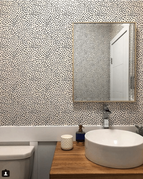 a powder room with light wood cabinetry. On top is a white sink bowl. Leo's Spots wallpaper manufactured by Milton & King and designed by Jillian Harris is on the wall and in the mirror reflecting the opposite wall.