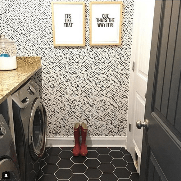 a laundry room with a dark metallic washer and dryer on the left and a white door on the right. the floor is a black geometric tile. on the far wall is wallpaper called Leo's Spots wallpaper manufactured by Milton & King and designed by Jillian Harris. On the wallpapered wall are two pictures framed that say "Its Like That" and "Cuz Thats The Way It Is". On the floor are red gumboots. 