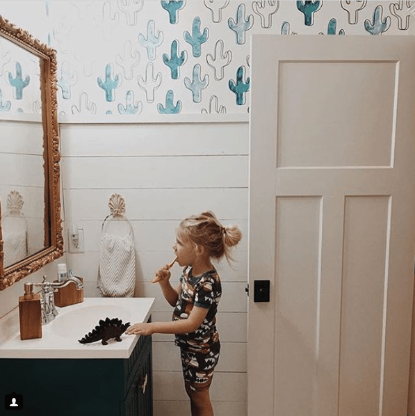 a young blonde long-haired boy brushing his teeth in front of a sink with white top on top of black cabinetry. He is looking into a mirror with a ornate gold frame. behind the boy is a wall with white horizontal timber on the bottom and on the top is blue and white Cactus Wallpaper by Milton & King.