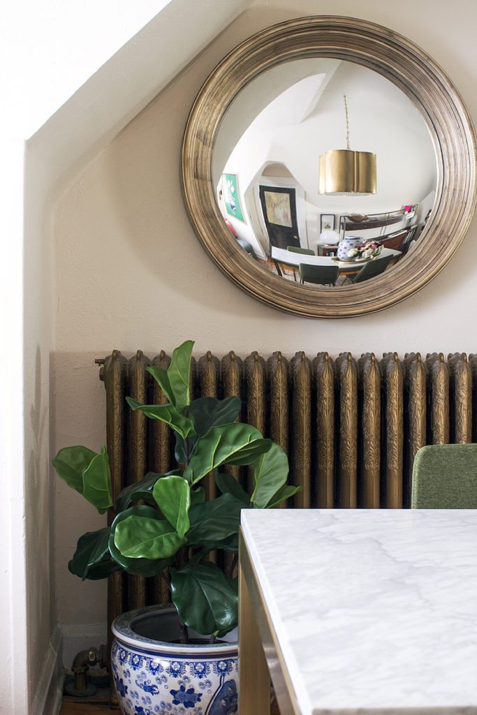 on the bottom left is a blue traditional planter with a large green leafy plant. On the wall is a circular convex mirror hanging above a brass wall heater. The marble top dining table is in the foreground