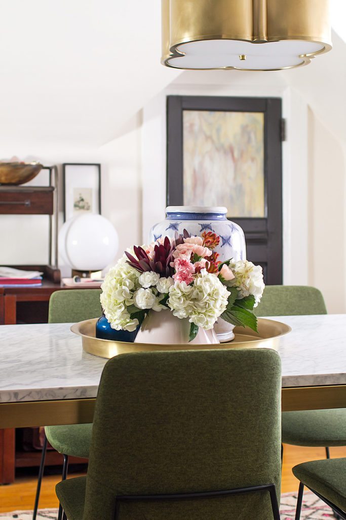 A close up shot of center piece of white, pink and red flowers on the table in the middle of the room. In the background is a blurred view of the door. In the foreground you can see the back of one of the olive green chairs 