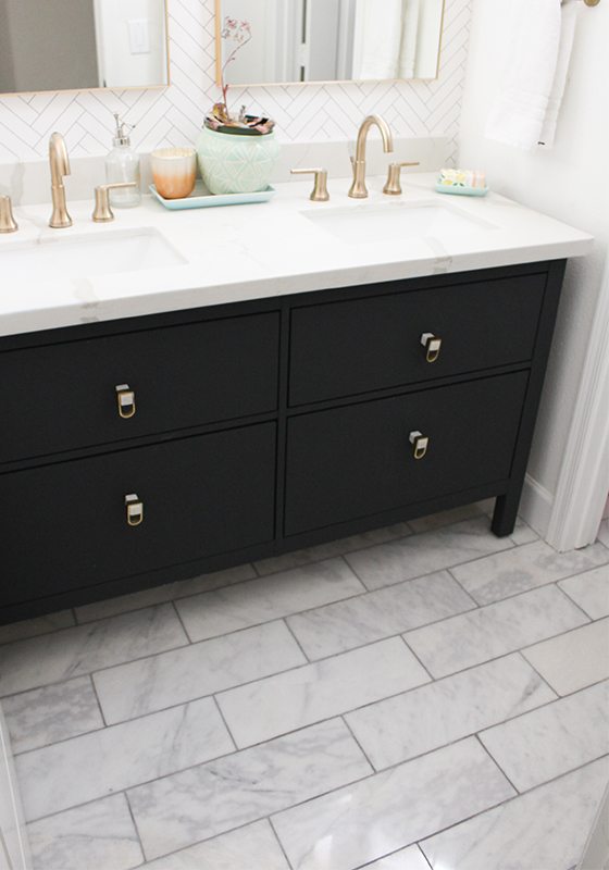 photo is focused on the dark cabinetry of the vanity which looks like it is painted black. It is on a marble tiled floor. On top is a his and hers sink with brass accents and the back wall has Tile Progress wallpaper from Milton & King