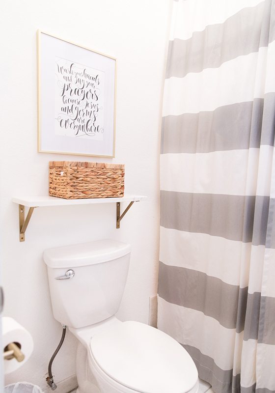 toilet with a shower on the right. The shower has a thick horizontally gray striped shower curtain. Above the toilet is a shelf with a whicker basket for storage and a picture above