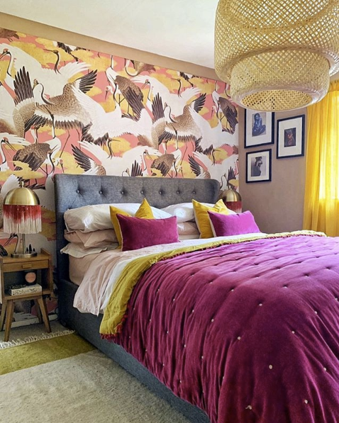 Pink Cranes Wallpaper behind a bed with a grey headboard. Gold and purple pillows are on the bed.