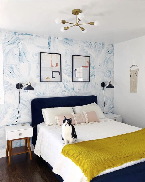 Shoreline blue wallpaper in guest bedroom behind bed with dark headboard. there is a cat on the bed looking at the camera and a golden throw blanket at the foot of the bed.