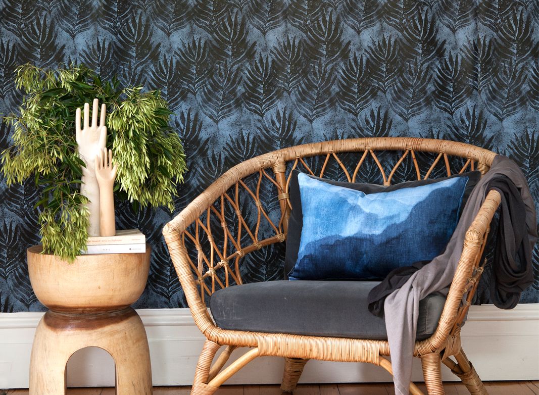 Shibori Leaf Wallpaper Design • Dark Blue Wallpaper behind a short wicker chair on the right and a green shrub on the left. In front of the green shrub are two carved wooden hands