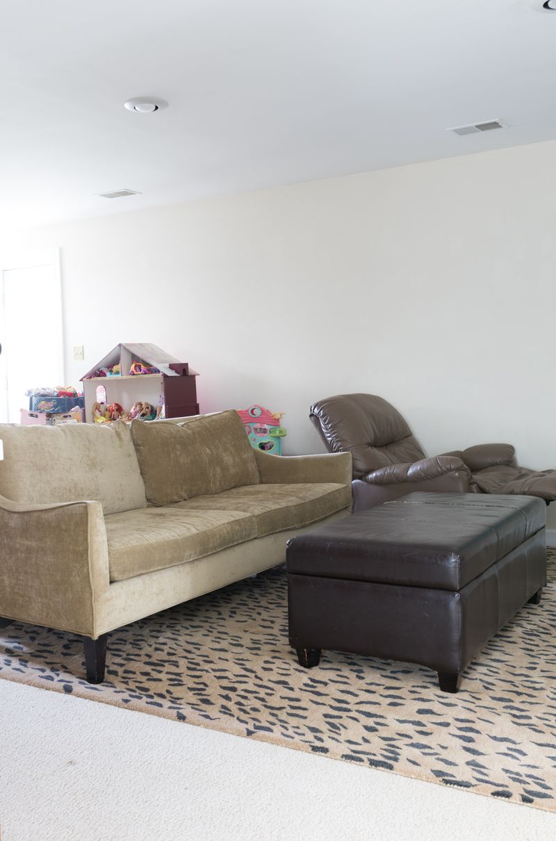 Jewel Marlow Basement Makeover • A beige couch is on a leopard rug with a dark brown leather ottoman and blank white walls in the background