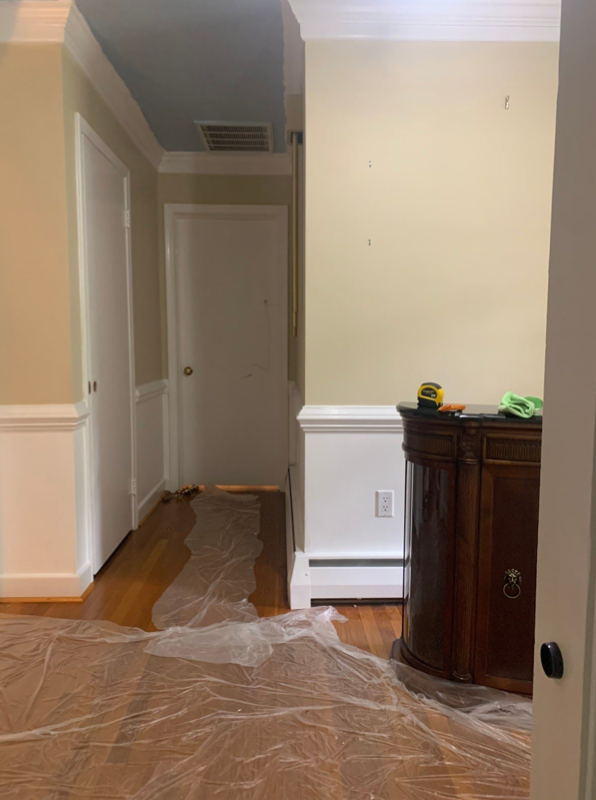 Picture of an entry way and a hallway with bare beige walls and white paint just below the chair rail. There is plastic on the floor in preparation for painting.